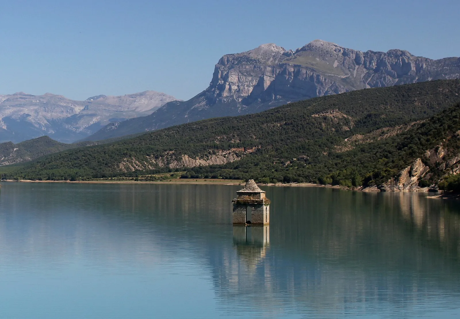 Torre de la iglesia de Mediano emergiendo de las aguas turquesa del embalse, con montañas al fondo